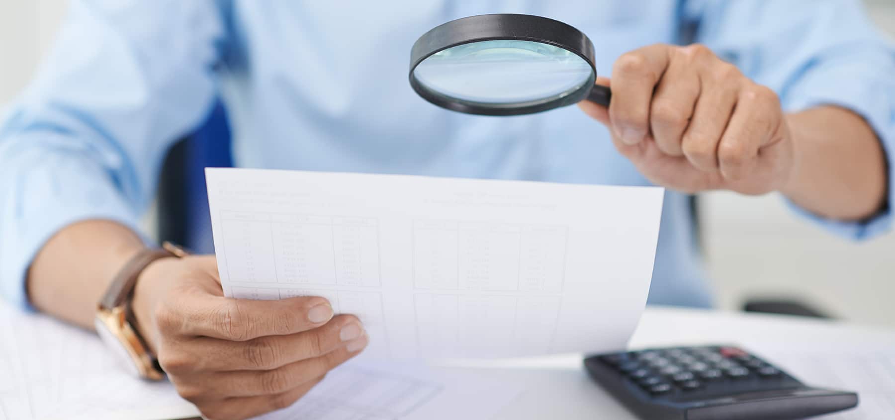Person looks at a paper with a magnifying glass with a calculator on table, looking for fraud in the business reports